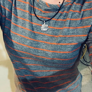 May include: A grey and orange striped long-sleeved shirt with a silver pendant necklace. The pendant is a round, detailed design.