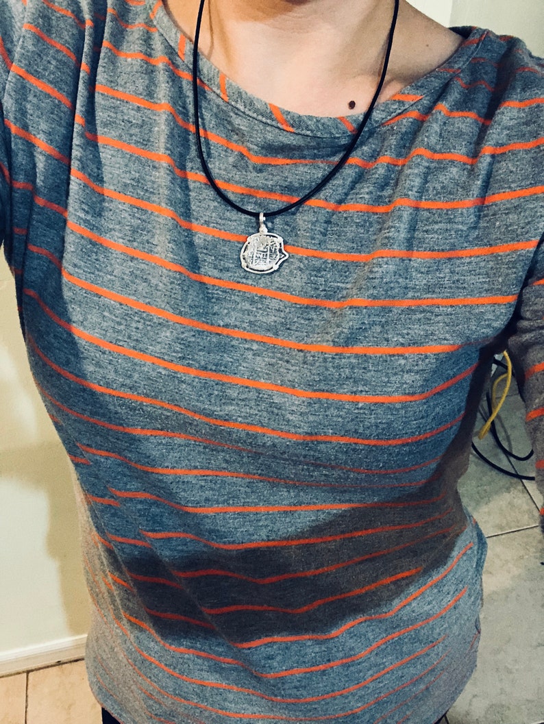 May include: A grey and orange striped long-sleeved shirt with a silver pendant necklace. The pendant is a round, detailed design.