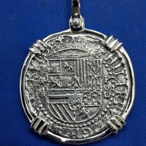 May include: A silver pendant with a coin-like design. The pendant features a detailed embossed coat of arms and text. It has a loop at the top for a chain. The pendant is set against a blue background.