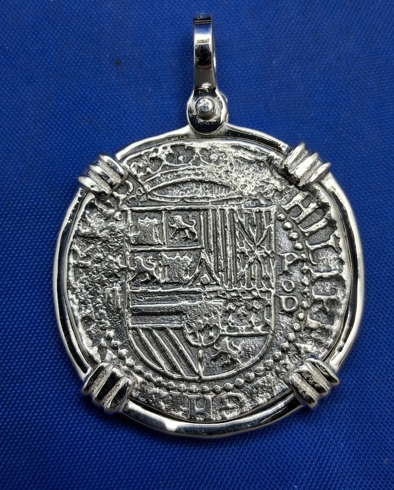 May include: A silver pendant with a coin-like design. The pendant features a detailed embossed coat of arms and text. It has a loop at the top for a chain. The pendant is set against a blue background.