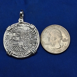 May include: A silver pendant with an ornate design, shown beside a U.S. quarter for scale. The pendant has a textured surface with detailed patterns and a loop for a chain. The quarter is approximately 2.4 cm in diameter.