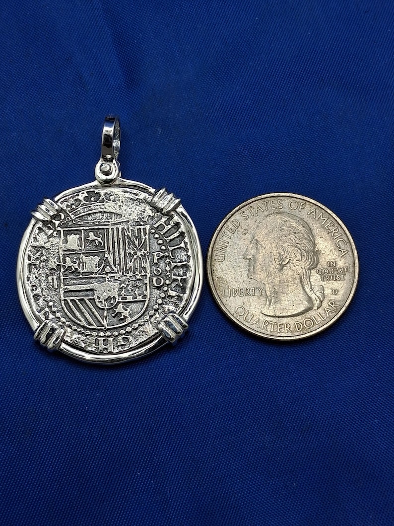 May include: A silver pendant with an ornate design, shown beside a U.S. quarter for scale. The pendant has a textured surface with detailed patterns and a loop for a chain. The quarter is approximately 2.4 cm in diameter.