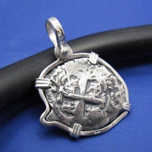 May include: Silver pendant featuring a replica of a Spanish colonial coin with a cross design and intricate details. The pendant is framed with a silver border and has a bail for attaching a chain.