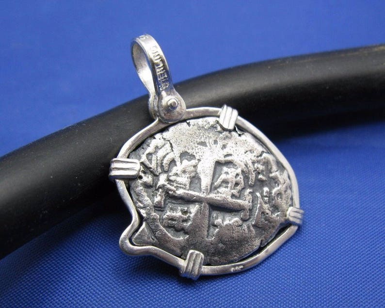 May include: Silver pendant featuring a replica of a Spanish colonial coin with a cross design and intricate details. The pendant is framed with a silver border and has a bail for attaching a chain.