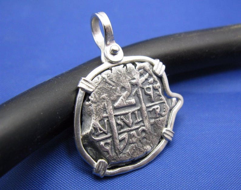 May include: Silver pendant featuring a replica of a Spanish colonial coin with a cross and inscription. The coin is set in a silver frame with a bail for attaching a chain.
