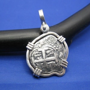 May include: Silver pendant featuring a replica of a Spanish eight reales coin, also known as a piece of eight. The coin is surrounded by a silver frame with decorative accents.