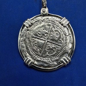 May include: A silver pendant with a coin-like design. The pendant features a detailed cross pattern in the centre, surrounded by intricate textures and a decorative border. It has a loop at the top for a chain.