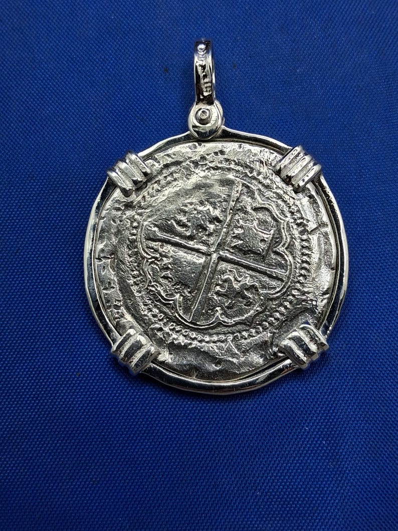May include: A silver pendant with a coin-like design. The pendant features a detailed cross pattern in the centre, surrounded by intricate textures and a decorative border. It has a loop at the top for a chain.