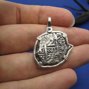May include: Silver pendant featuring a replica of a Spanish eight reales coin, also known as a piece of eight. The coin features a cross and the inscription "+ HISPANIARVM +" on the front.
