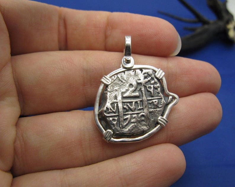 May include: Silver pendant featuring a replica of a Spanish eight reales coin, also known as a piece of eight. The coin features a cross and the inscription "+ HISPANIARVM +" on the front.