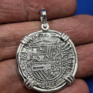 May include: A silver coin pendant with a detailed design, including a crown and shield. The coin has a textured surface and a loop for a chain. The pendant is held in a hand against a blue backdrop.