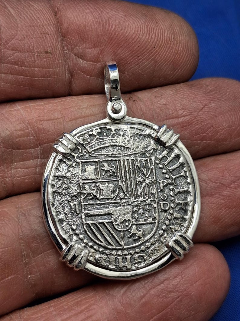 May include: A silver coin pendant with a detailed design, including a crown and shield. The coin has a textured surface and a loop for a chain. The pendant is held in a hand against a blue backdrop.