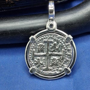 May include: A silver pendant with a textured coin design and a cross symbol. The pendant has a circular frame with four decorative accents and a loop for a chain. The background is a blue fabric.