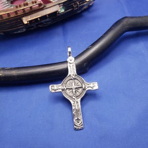 May include: A silver pendant in the shape of a cross with a compass design in the center. The cross is detailed with intricate carvings and has a loop at the top for attaching a chain.