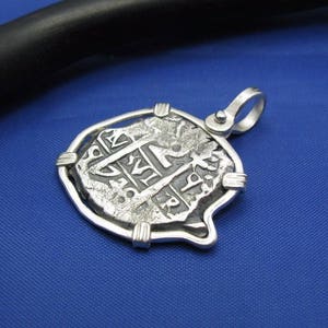 May include: A silver pendant featuring a replica of a historic Spanish coin, encased in a protective silver frame. The coin has intricate details and lettering, including the year "1718".