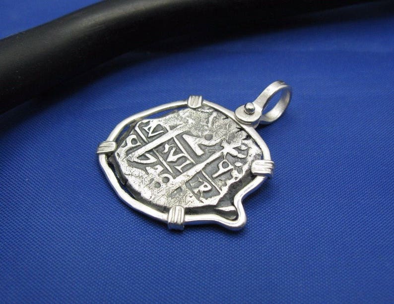 May include: A silver pendant featuring a replica of a historic Spanish coin, encased in a protective silver frame. The coin has intricate details and lettering, including the year "1718".