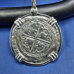 May include: Silver pendant with a detailed coin design, featuring a cross and intricate patterns. The pendant has a loop for a chain and four decorative clasps around the edge. The coin is set against a blue background.