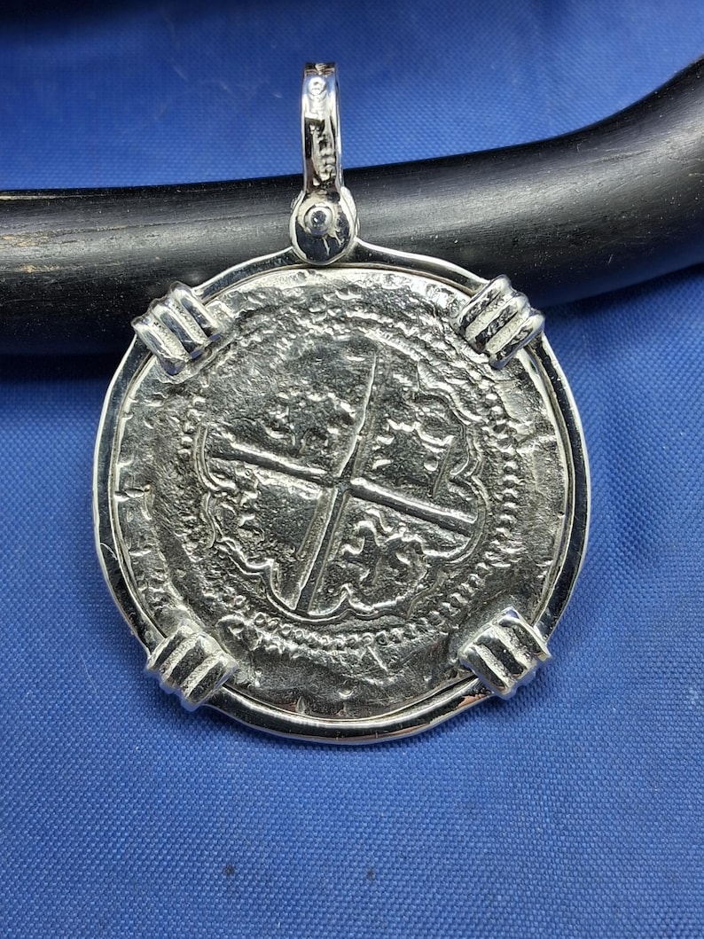 May include: Silver pendant with a detailed coin design, featuring a cross and intricate patterns. The pendant has a loop for a chain and four decorative clasps around the edge. The coin is set against a blue background.