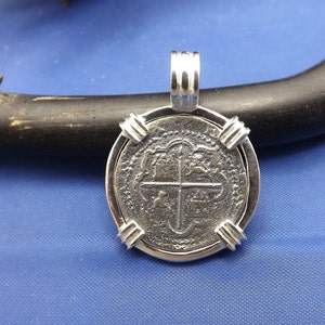 May include: Silver pendant featuring a replica of a Spanish colonial coin with a cross design. The coin is framed with a silver bezel and has a bail for attaching to a chain.