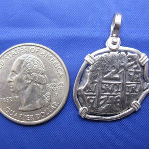 May include: A silver replica of a Spanish 2 reales coin, also known as a "piece of eight", is shown on a blue background. The coin is mounted on a silver pendant with a loop for a chain. A U.S. quarter is shown for size comparison.