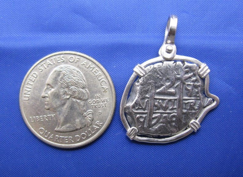 May include: A silver replica of a Spanish 2 reales coin, also known as a "piece of eight", is shown on a blue background. The coin is mounted on a silver pendant with a loop for a chain. A U.S. quarter is shown for size comparison.
