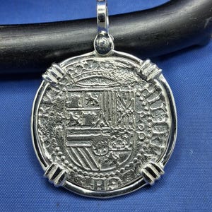 May include: Silver pendant with a coin-like design. The pendant has a detailed central emblem and a textured border. It includes a loop at the top for a chain. The design is highly detailed.