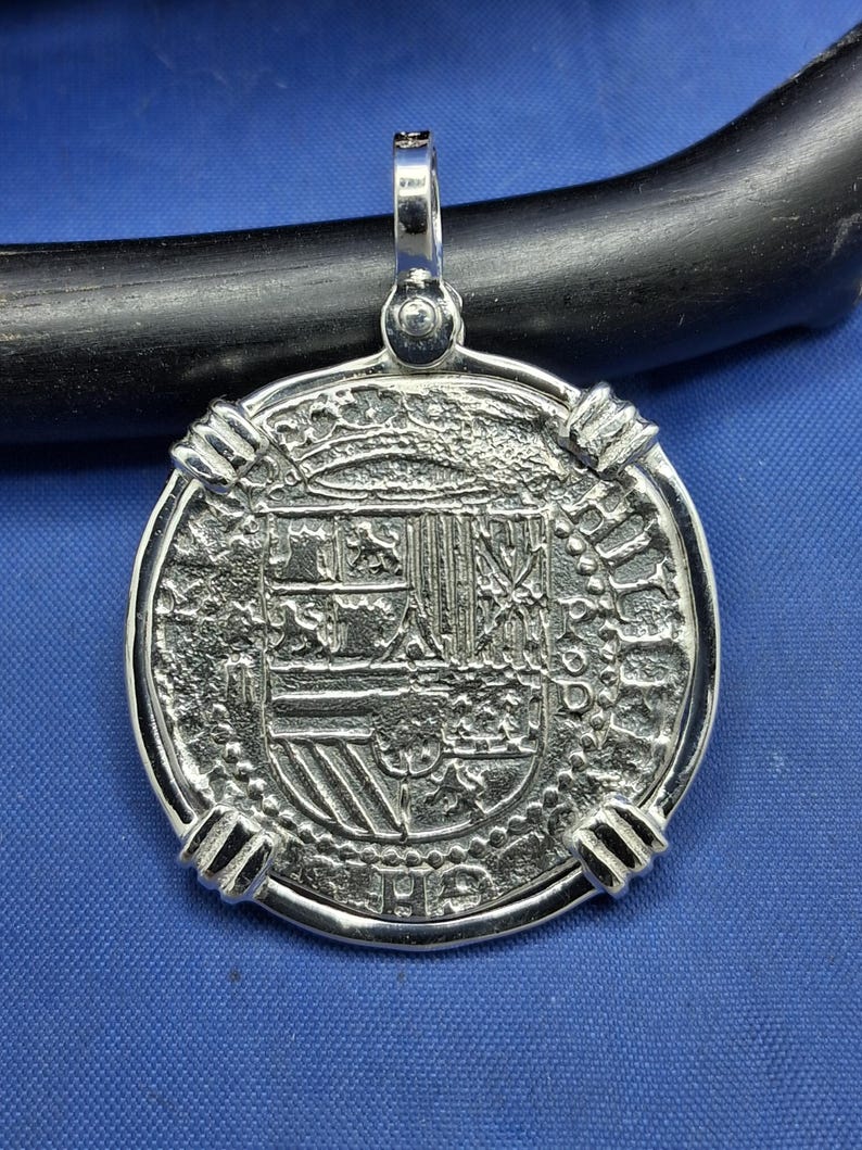 May include: Silver pendant with a coin-like design. The pendant has a detailed central emblem and a textured border. It includes a loop at the top for a chain. The design is highly detailed.