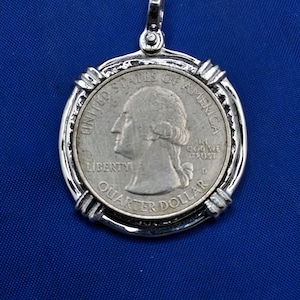 May include: A silver pendant featuring a United States quarter coin. The coin is set within a decorative silver frame with a loop for a chain. The coin displays the profile of George Washington and the words "Liberty" and "Quarter Dollar."