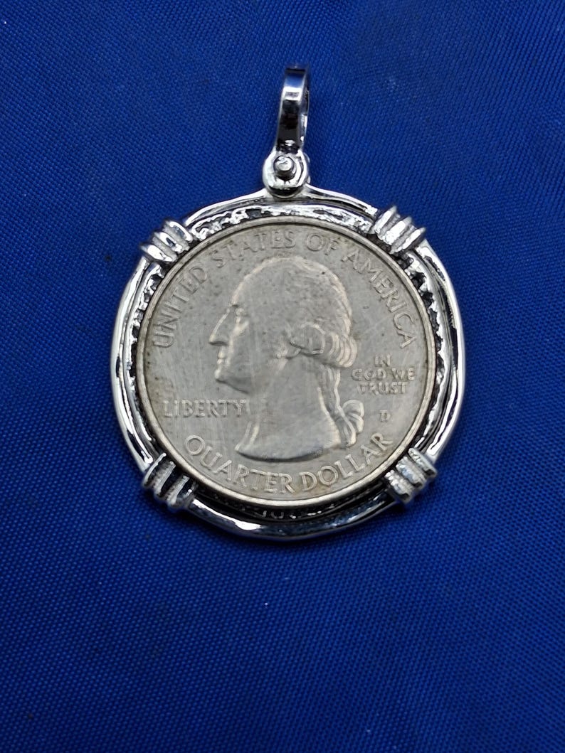 May include: A silver pendant featuring a United States quarter coin. The coin is set within a decorative silver frame with a loop for a chain. The coin displays the profile of George Washington and the words "Liberty" and "Quarter Dollar."