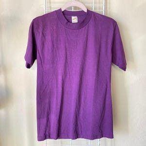 May include: A solid purple short-sleeve t-shirt hanging on a white wire rack. The shirt has a crew neck and a small tag at the neckline. The fabric appears to be a soft, lightweight material. The shirt is a vibrant shade of purple.