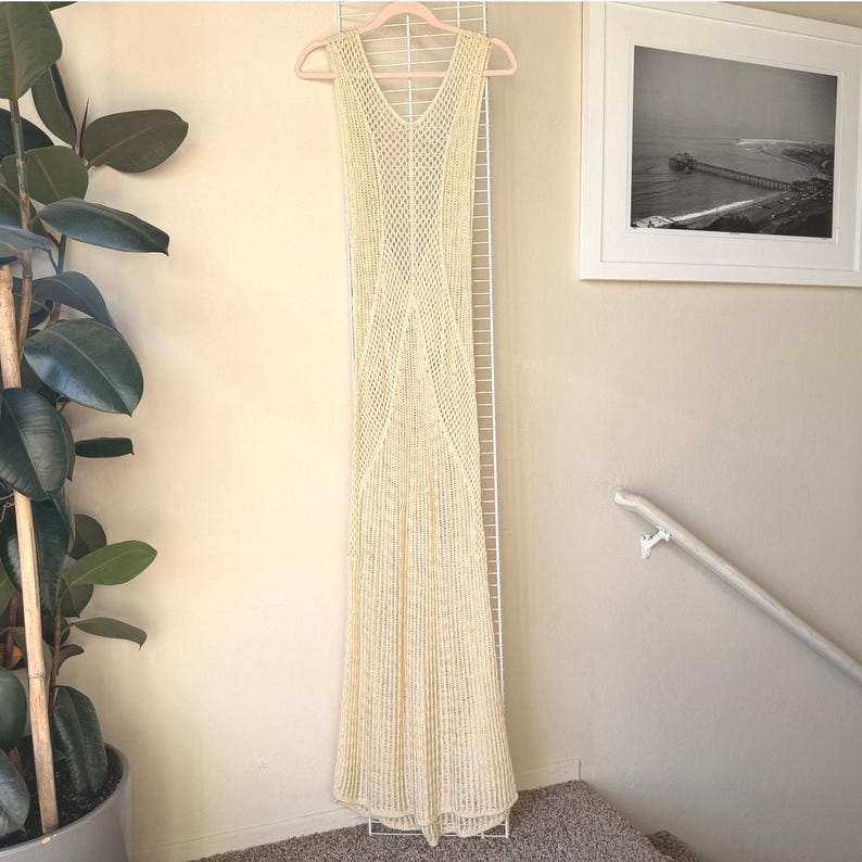 K&ouml;nnte beinhalten: Ein cremefarbenes, &auml;rmelloses, bodenlanges Kleid mit V-Ausschnitt. Das Kleid besteht aus geh&auml;keltem Material mit einem figurbetonten Oberteil und einem ausgestellten Rock. Es h&auml;ngt an einem rosa Kleiderb&uuml;gel vor einem wei&szlig;en Gitter.