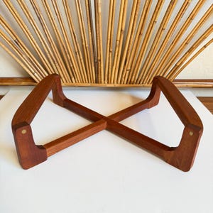 Puede incluir: Un soporte de madera en forma de X con un acabado marrón rojizo. El soporte tiene un diseño moderno de mediados de siglo, con patas anguladas y una construcción robusta. El soporte mide aproximadamente 30 cm de largo.