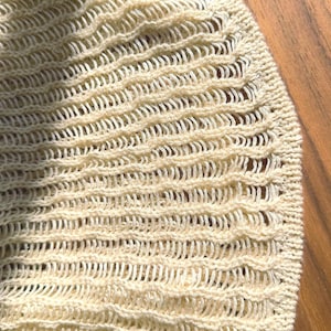 K&ouml;nnte beinhalten: Nahaufnahme eines cremefarbenen Strickstoffs mit einem strukturierten Lochmuster. Das Strickmuster weist horizontale Reihen von gebogenen, wellenartigen Formen auf, die ein zartes und luftiges Aussehen erzeugen. Die Textur des Stoffes ist sichtbar und unterstreicht die Handwerkskunst.