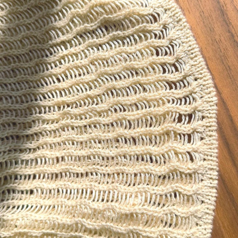 K&ouml;nnte beinhalten: Nahaufnahme eines cremefarbenen Strickstoffs mit einem strukturierten Lochmuster. Das Strickmuster weist horizontale Reihen von gebogenen, wellenartigen Formen auf, die ein zartes und luftiges Aussehen erzeugen. Die Textur des Stoffes ist sichtbar und unterstreicht die Handwerkskunst.