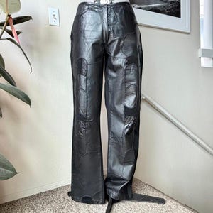 May include: Black leather pants featuring a patchwork design with multiple stitched panels. The trousers have a high-waisted fit, a front zipper closure, and a straight-leg silhouette. The surface of the leather shows varied textures and seams.