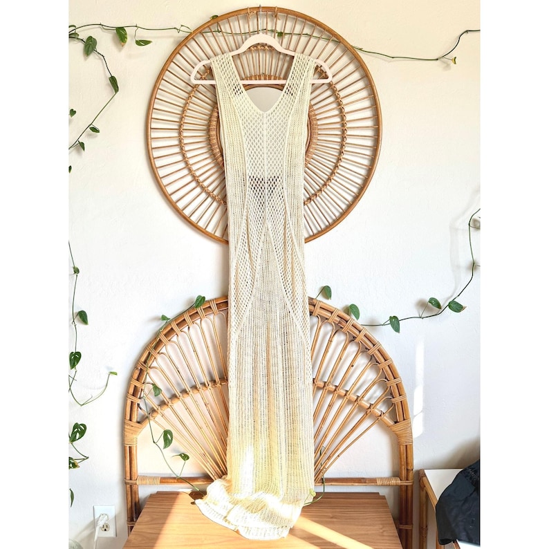 K&ouml;nnte beinhalten: Ein &auml;rmelloses, cremefarbenes, bodenlanges Kleid mit V-Ausschnitt und offenem Strickdesign. Das Kleid h&auml;ngt an einem wei&szlig;en Kleiderb&uuml;gel vor einem Hintergrund aus Wanddekoration aus Weide und einem Holztisch.