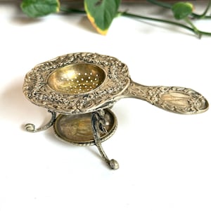 May include: A silver-toned metal tea strainer with an ornate, embossed floral pattern on the handle and rim. The circular bowl features small drainage holes and rests on a matching three-legged stand with a small circular drip tray underneath.