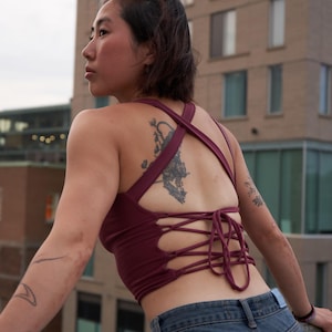 Puede incluir: Una mujer con un top corto de color granate con la espalda atada con cordones y pantalones vaqueros azules. El top tiene un diseño cruzado en la espalda y está atado a la cintura.