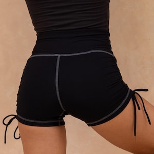 May include: Black athletic shorts with drawstring ties and ruching detail. The shorts have a high waist and are paired with a black tank top. The shorts are designed for fitness activities.