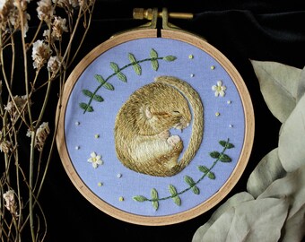 May include: A hand-stitched embroidery hoop featuring a sleeping dormouse curled up on a light blue background. The hoop is decorated with green leaves and white flowers.