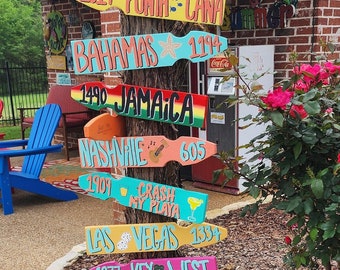 May include: A wooden signpost with colorful directional signs pointing to various destinations, including Punta Cana, Bahamas, Jamaica, Nashville, Crash My Playa, Las Vegas, and Key West. The signs are painted in bright colors and feature the destination names and distances.
