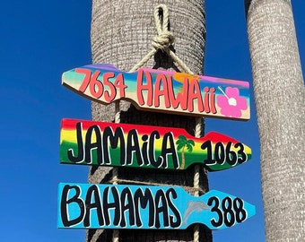 May include: Hand-painted wooden directional signs with the destinations Hawaii, Jamaica, and Bahamas. Each sign is a different color and features the destination name, a number, and a small graphic. The signs are hanging from a rope.