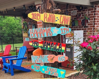 May include: A wooden signpost with colorful directional signs pointing to various destinations, including Punta Cana, Bahamas, Jamaica, Nashville, Crash My Playa, Las Vegas, and Key West. The signs are painted in bright colors with stylized text and distances.