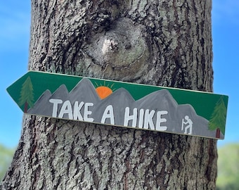 May include: Wooden arrow-shaped sign with the text "TAKE A HIKE" in white. The sign features a green border with mountain and sun illustrations. It is attached to a tree trunk, suggesting outdoor or hiking themes.