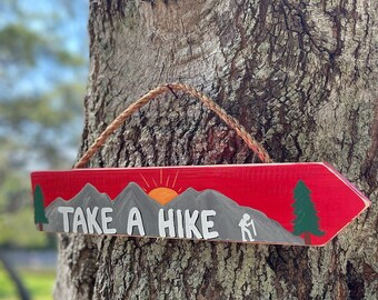 May include: Wooden arrow-shaped sign with a red background and a mountain and sun design. The sign reads "TAKE A HIKE" in white letters, with a hiker silhouette. A rope is attached for hanging.
