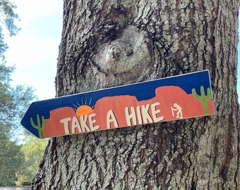 May include: Wooden sign with the text "TAKE A HIKE" in a desert landscape design. The sign features a blue sky, red rock formations, a setting sun, a cactus, and a hiker silhouette. The sign is attached to a tree trunk.
