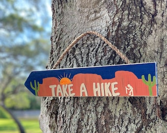 May include: Wooden arrow-shaped sign with the text "TAKE A HIKE" in a desert landscape design. The sign features a blue sky, orange mountains, a setting sun, and a hiker silhouette. A cactus is on the left side.