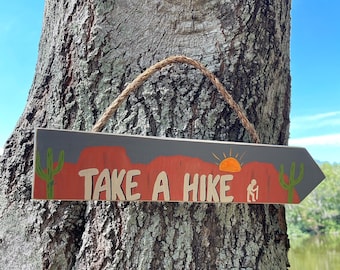 May include: Wooden arrow-shaped sign with the text "TAKE A HIKE" in a desert landscape design. The sign features a gray background, red mountains, a setting sun, and two green cacti. A rope is attached for hanging.