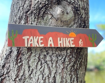 May include: Wooden arrow-shaped sign with the words "TAKE A HIKE" in white, featuring a desert landscape design. The sign includes a red-orange mountain range, a yellow sun, and green cacti. It is attached to a tree.