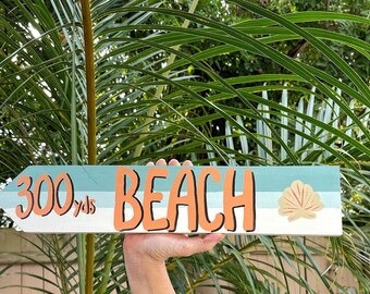 May include: Wooden beach sign with an arrow shape. The sign reads "300 yds BEACH" in orange lettering. A seashell graphic is on the right. The sign is painted with a blue, white, and orange color scheme.